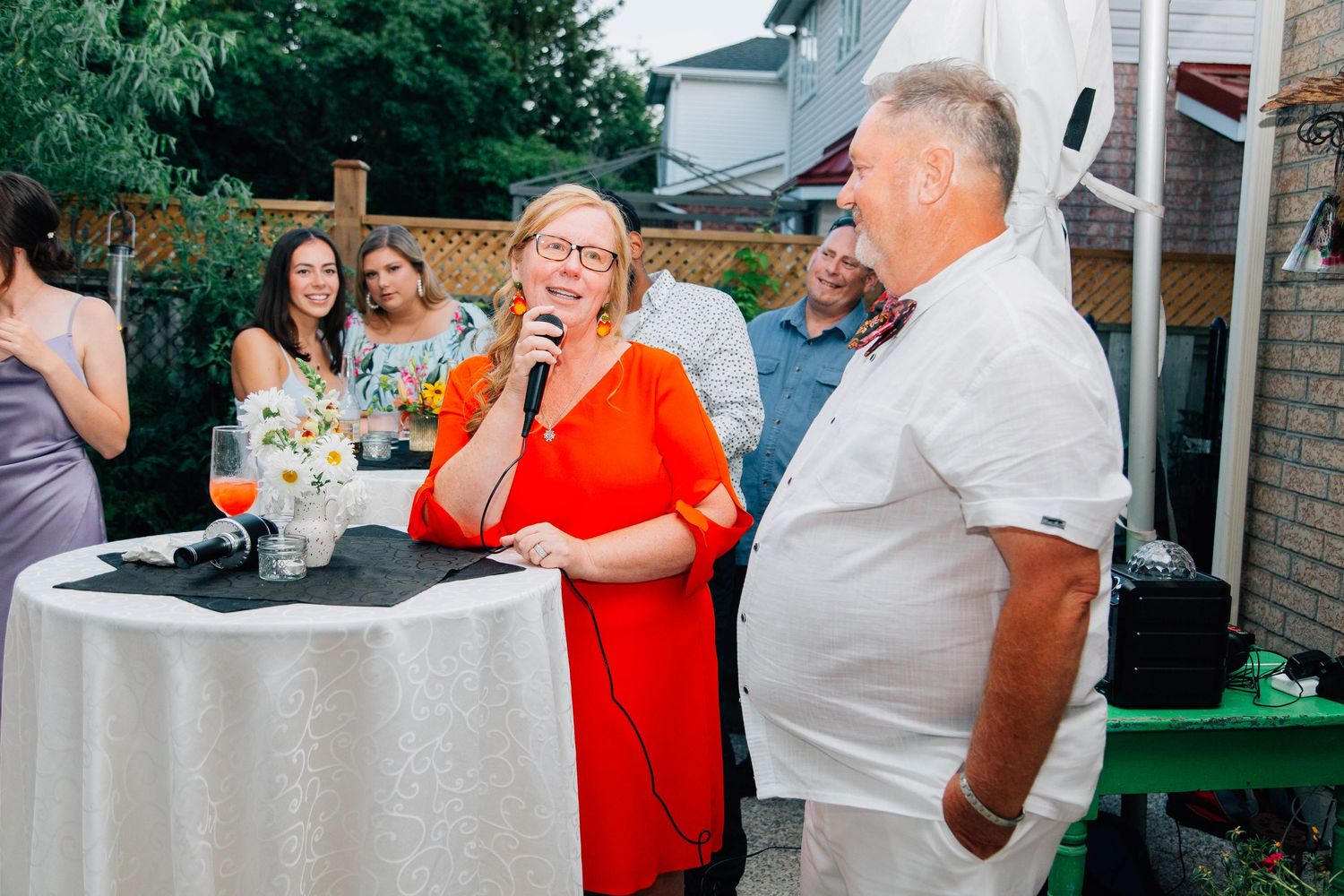 Woman in red dress speaks into microphone at outdoor wedding reception while guests look on.