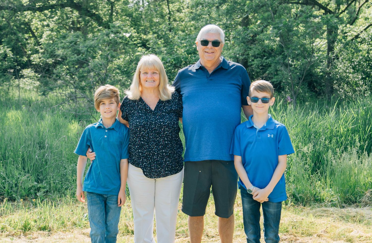 Grandparents posing outdoors with two young boys in matching blue polo shirts in a green natural setting.