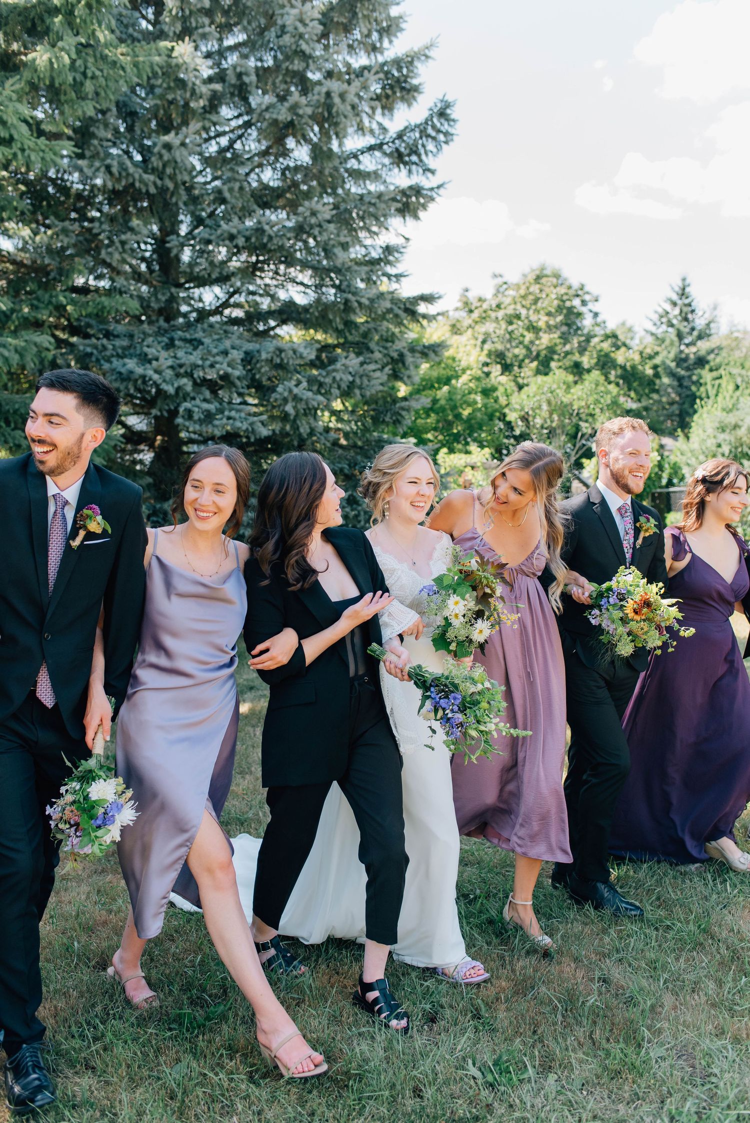 Bride in white gown walking with bridesmaids in purple and black outfits carrying colorful bouquets outdoors.