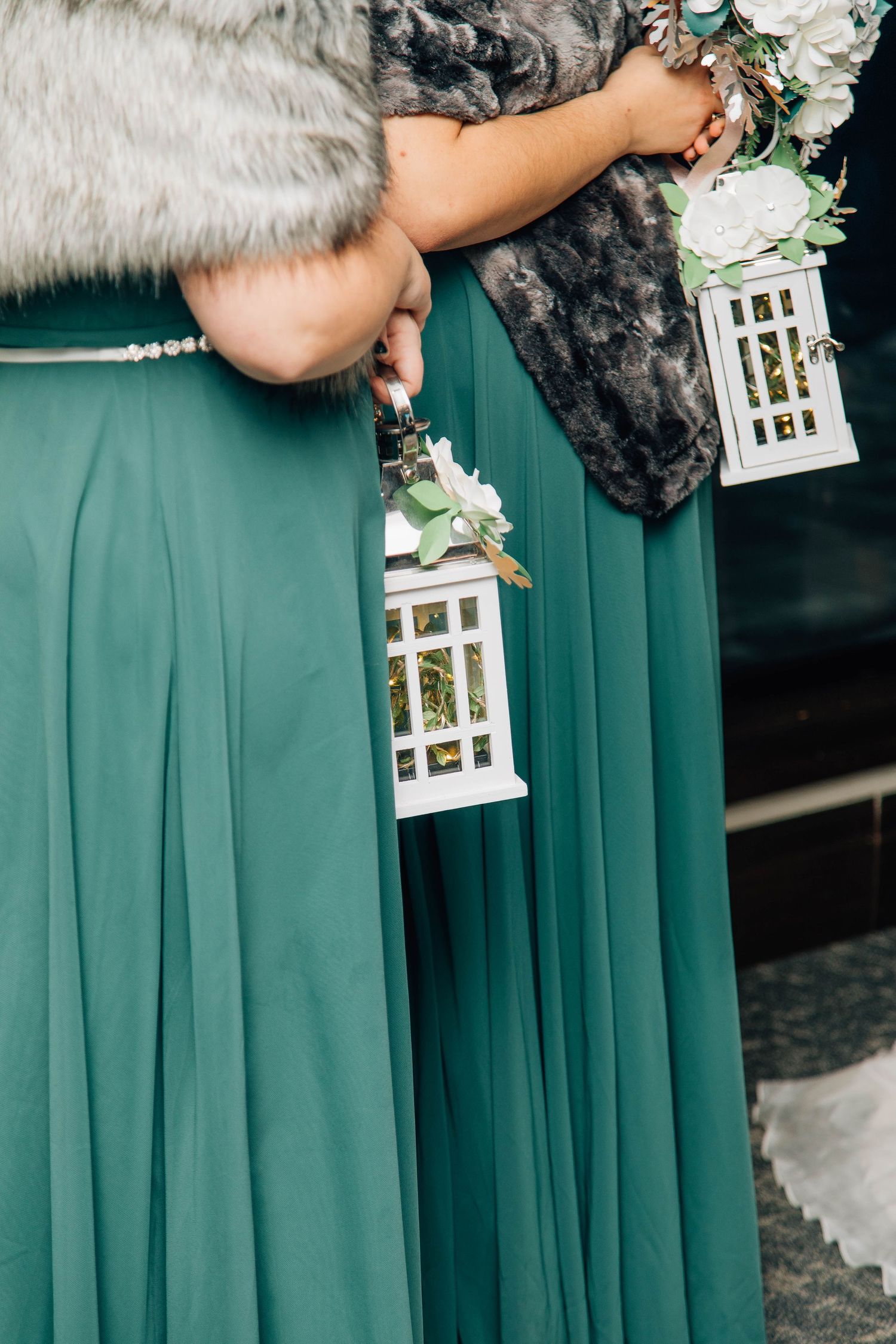 Bridesmaids in emerald green gowns with fur wraps hold white decorative lanterns and floral bouquets.