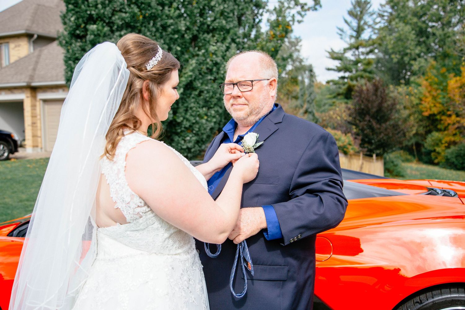 Bride pinning boutonniere on father's navy suit lapel beside red sports car outdoors.