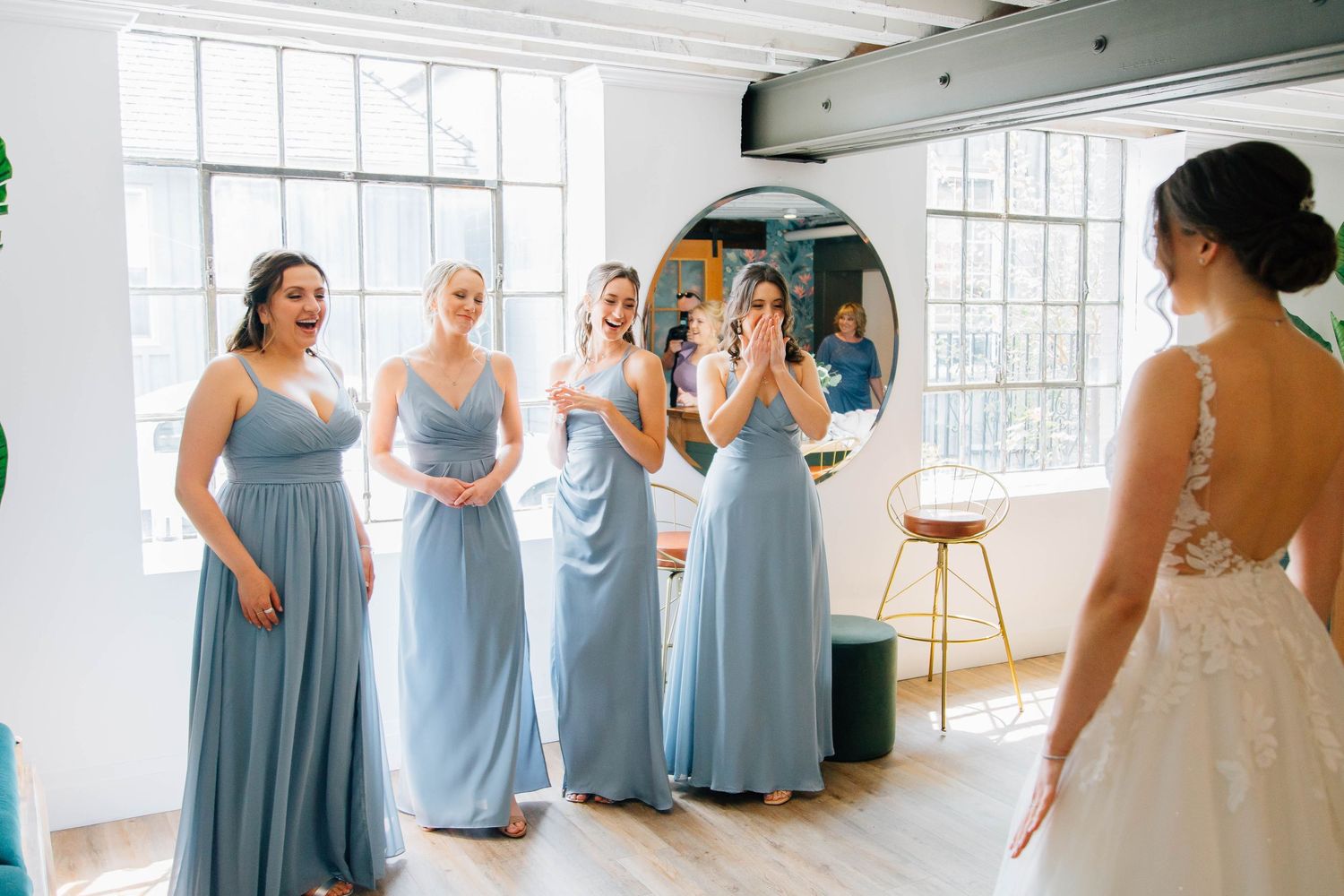 Bridesmaids in dusty blue dresses gasp in awe at the bride in her lace wedding gown at the Timber Barn in St. Jacobs Ontario.
