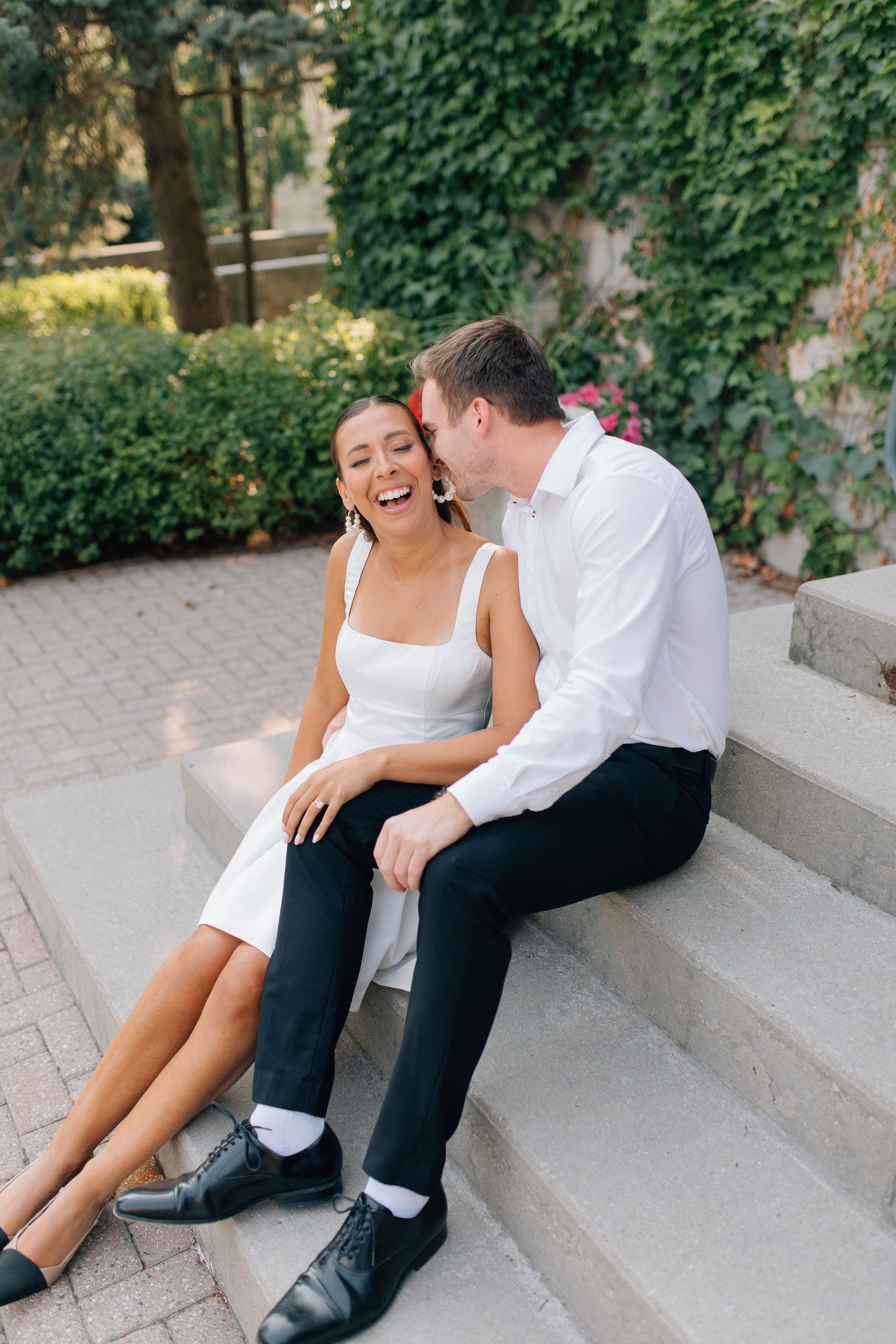 Laughing couple sits on stone steps as groom whispers in brides ear outdoors.