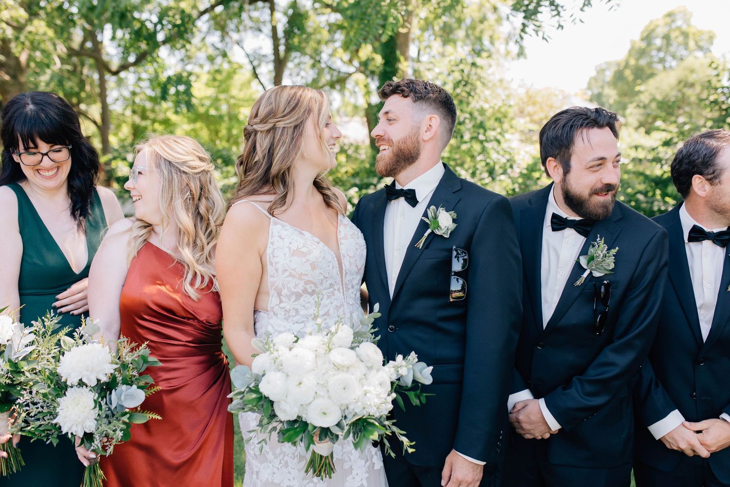 Bride holding white bouquet laughing with groom and wedding party outdoors.
