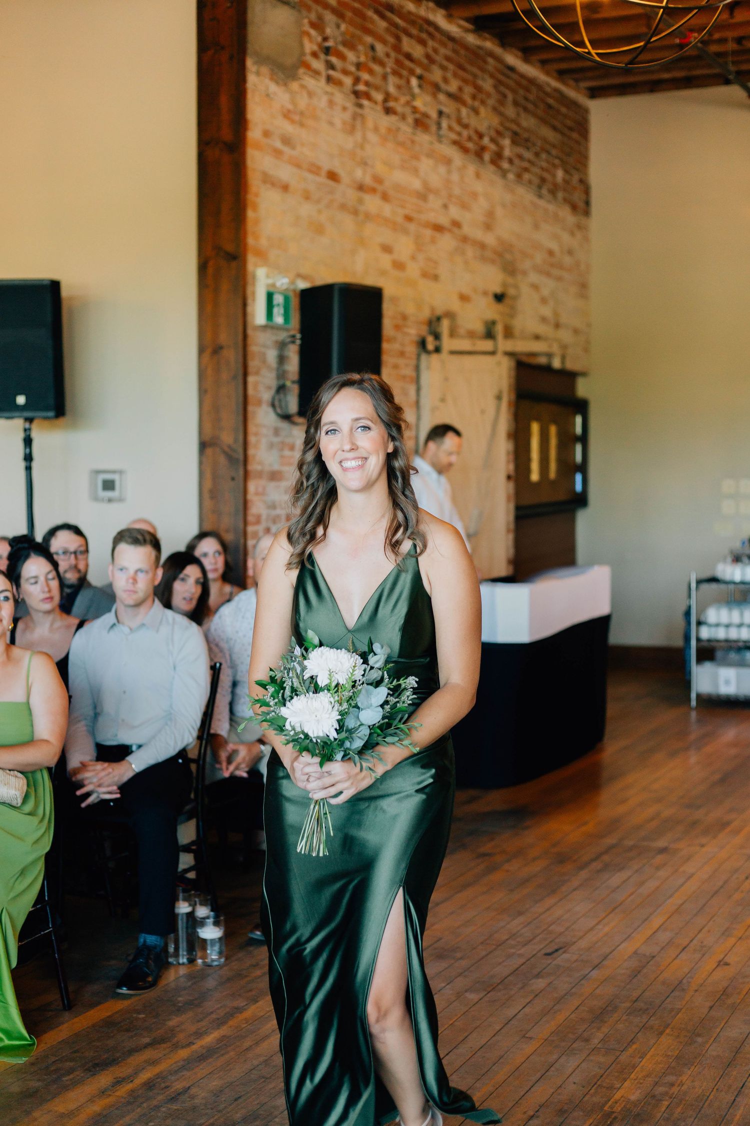 Smiling bridesmaid in emerald green satin gown carries white bouquet down aisle at indoor wedding ceremony.