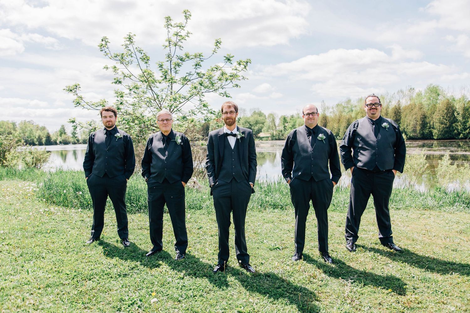 Five groomsmen in black suits standing casually on grass by a scenic pond on a wedding day.