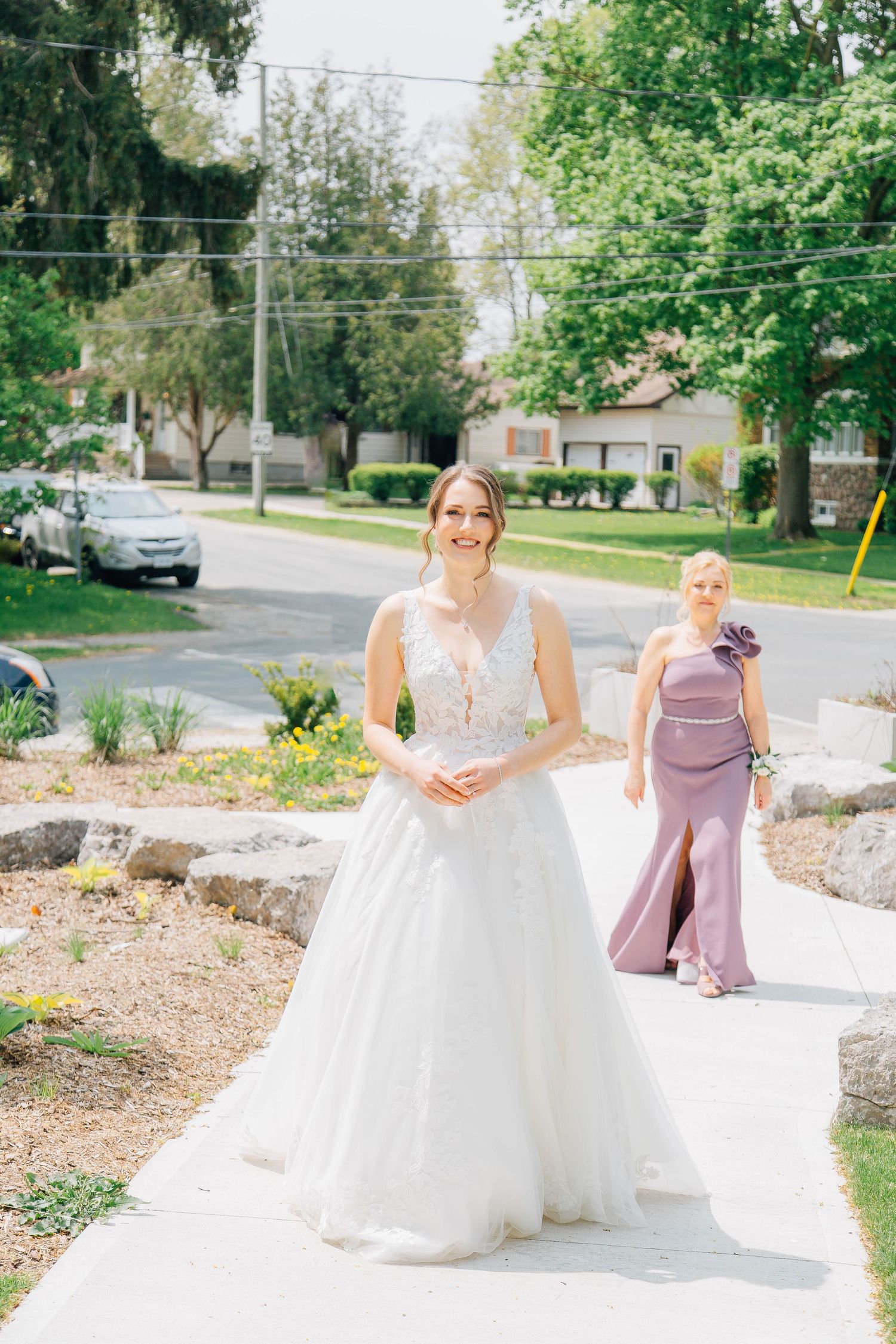 Radiant bride in lace ball gown walks pathway with bridesmaid in lavender dress on sunny wedding day.