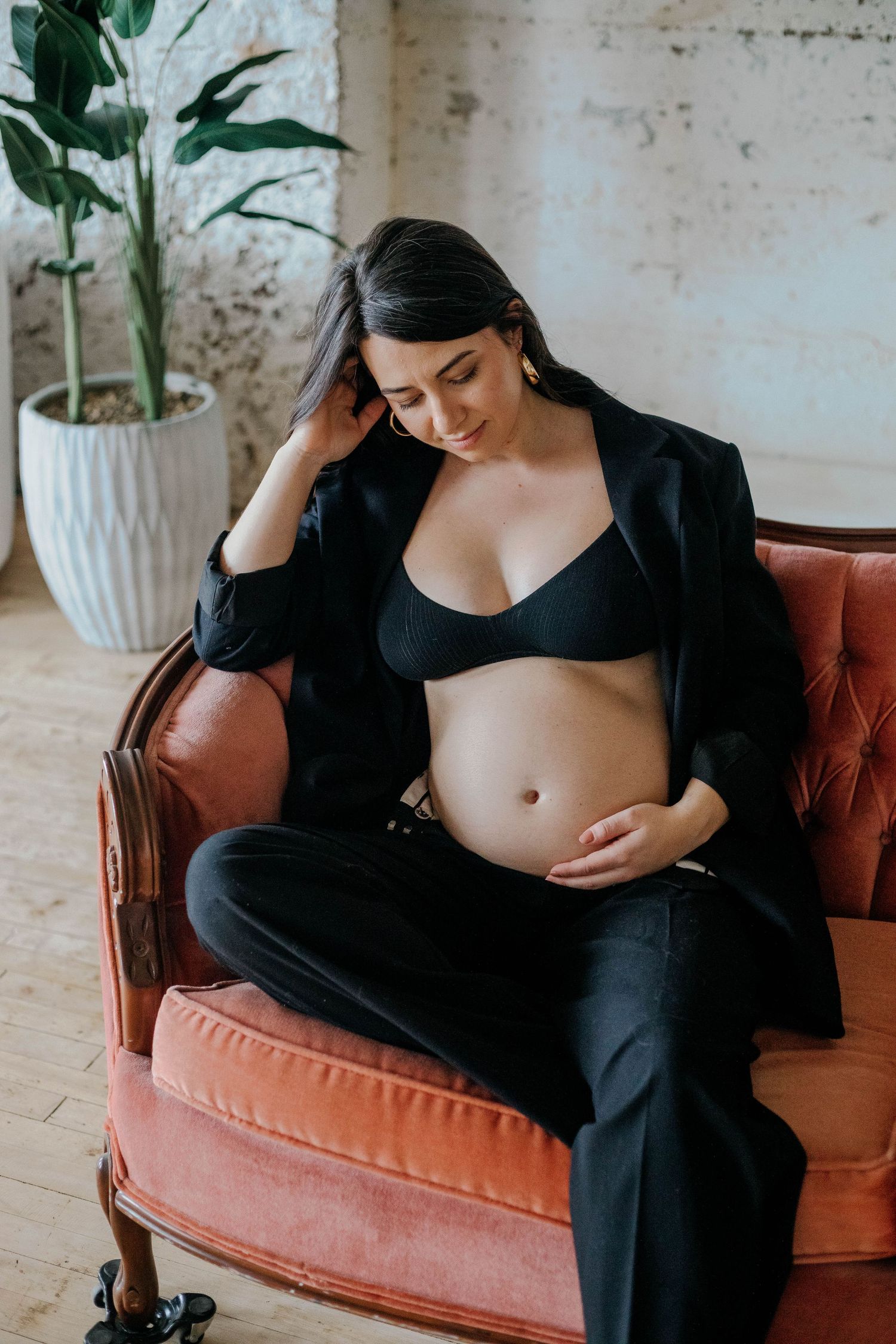 Pregnant woman in black blazer relaxing in a vintage coral armchair, gently touching her bare baby bump.
