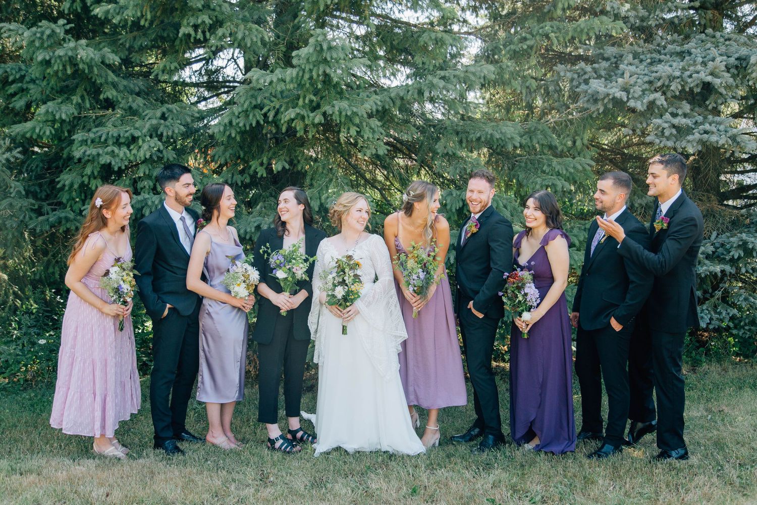 A wedding party in purple and black attire laughing together on a green lawn.