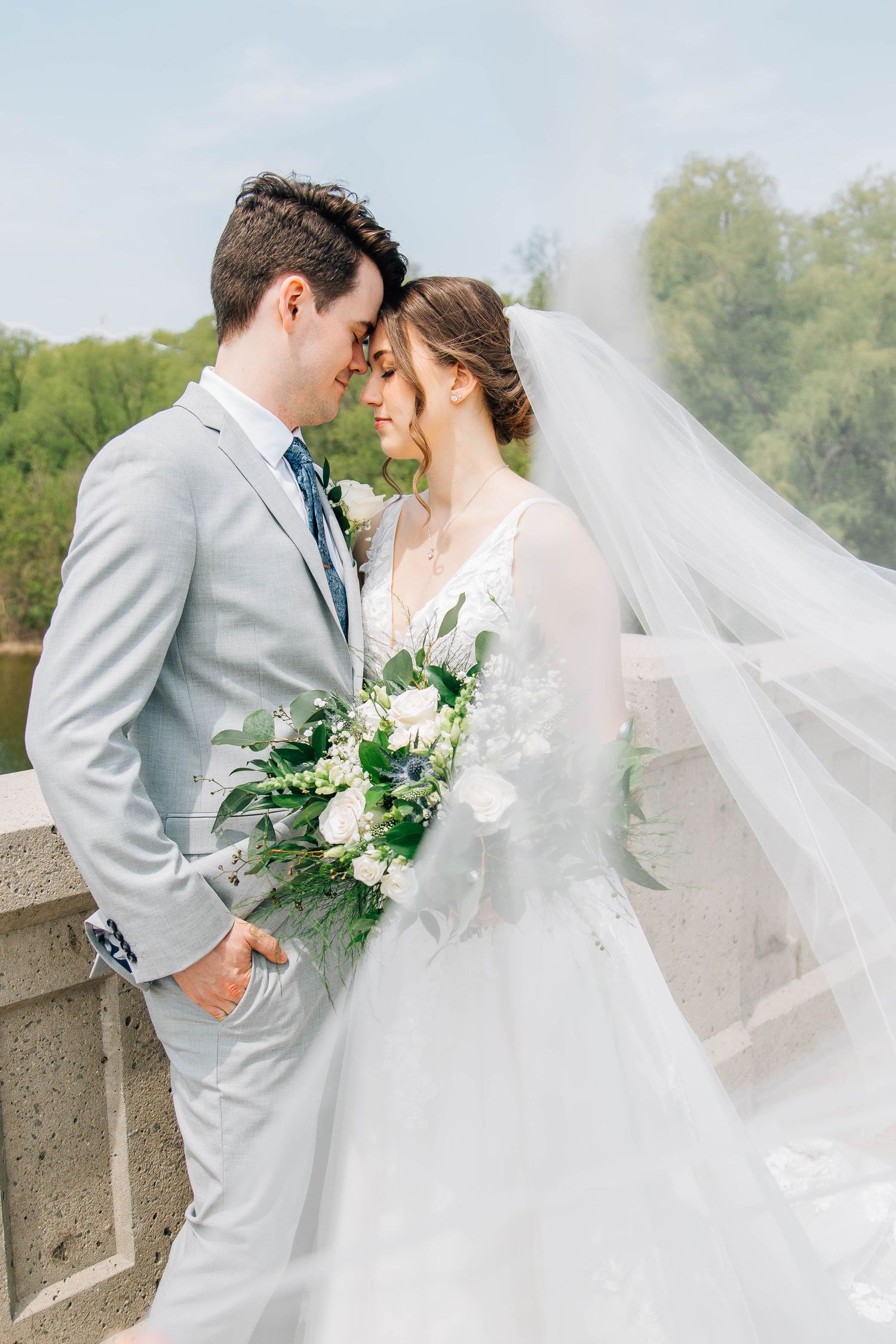 Romantic bride and groom touching foreheads on a bridge, brides veil flowing with a white bouquet.