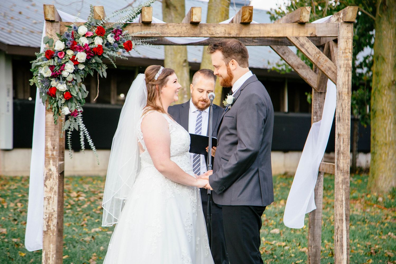Bride and groom exchange vows under a rustic wooden arch with red floral decor.