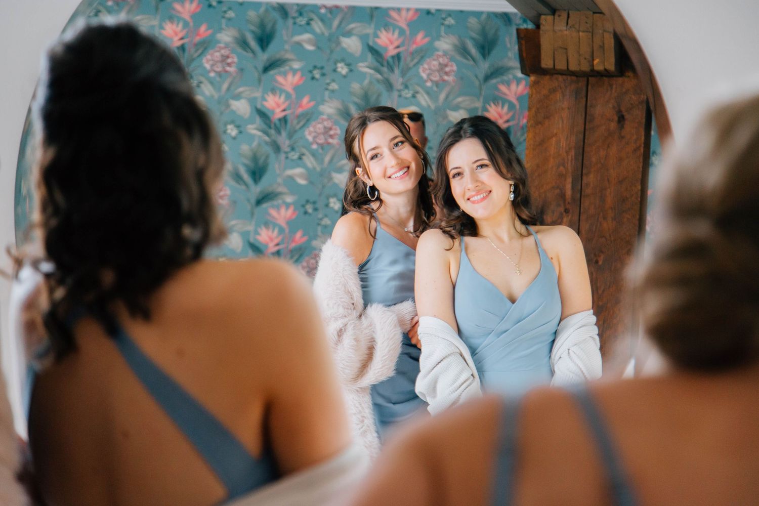 Two smiling bridesmaids in dusty blue dresses sit together during wedding preparations.