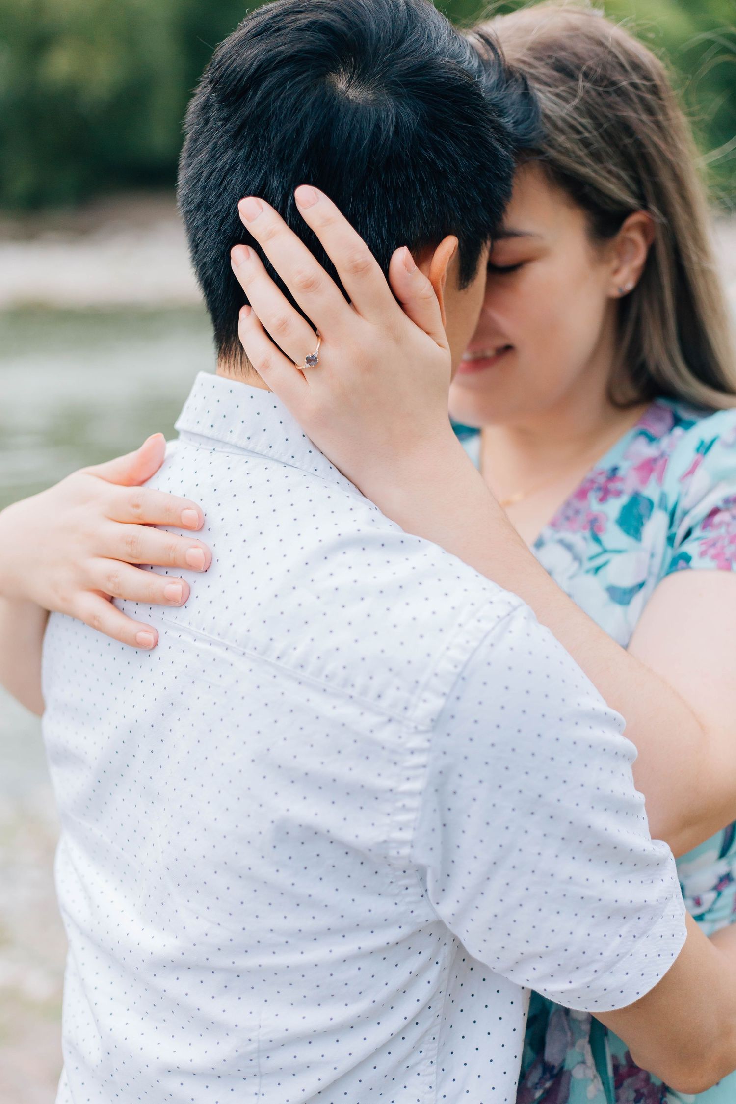 Close-up of couple embracing near water, woman showing engagement ring while hugging man.