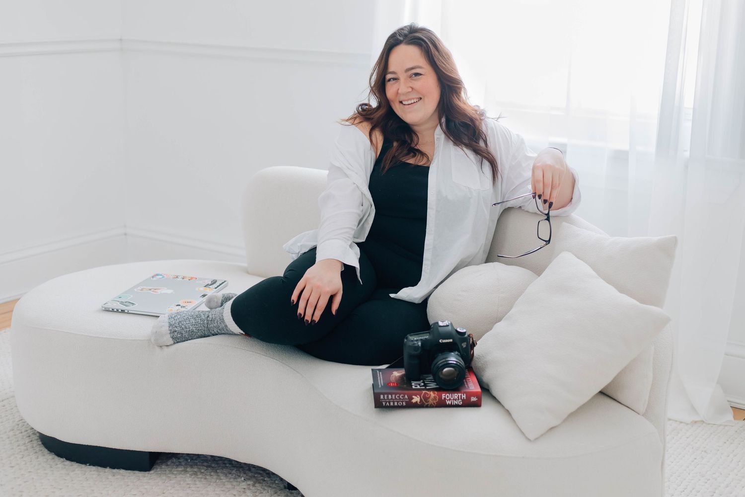Smiling female photographer sitting on a white curved sofa with her camera in a bright white studio.