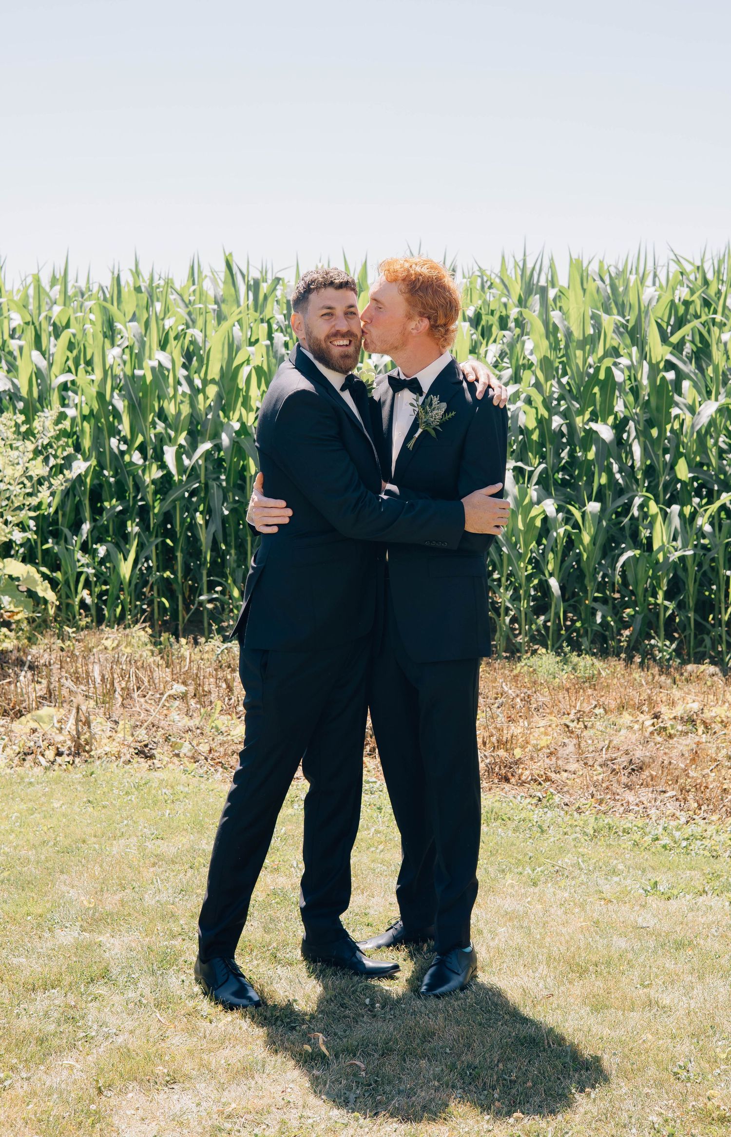 Two grooms in black tuxedos sharing a tender moment in front of a green cornfield on their wedding day.