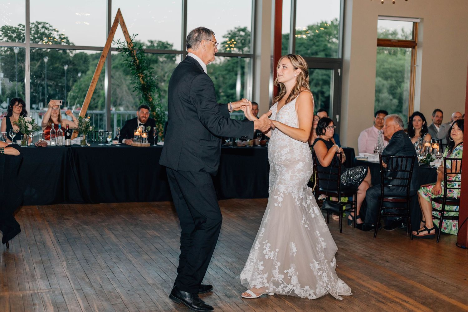 Bride in floral lace gown dances with father during father-daughter dance at wedding reception.