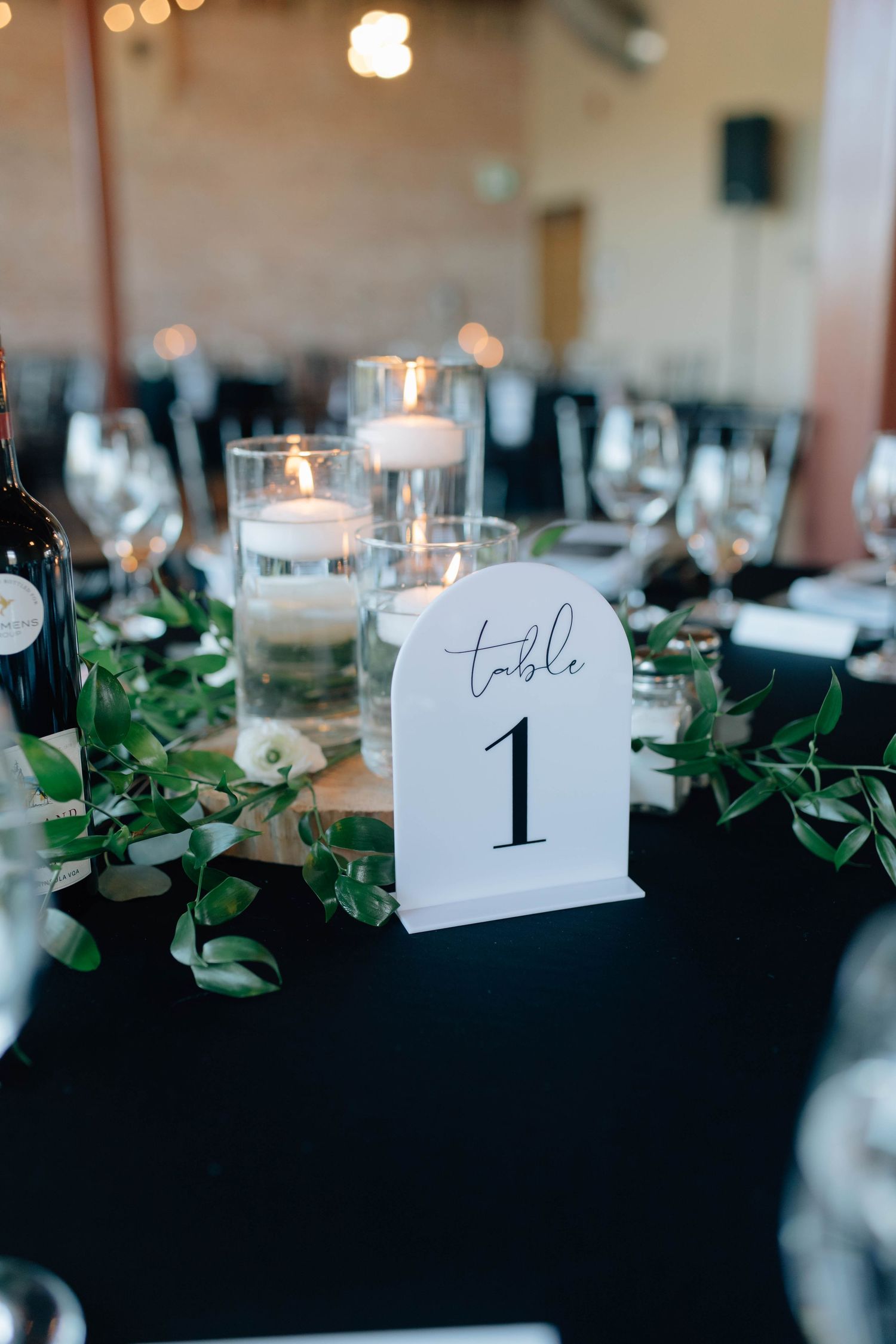Wedding reception table with arch-shaped Table 1 sign, floating candles, greenery runner, and wine glasses on black linen.