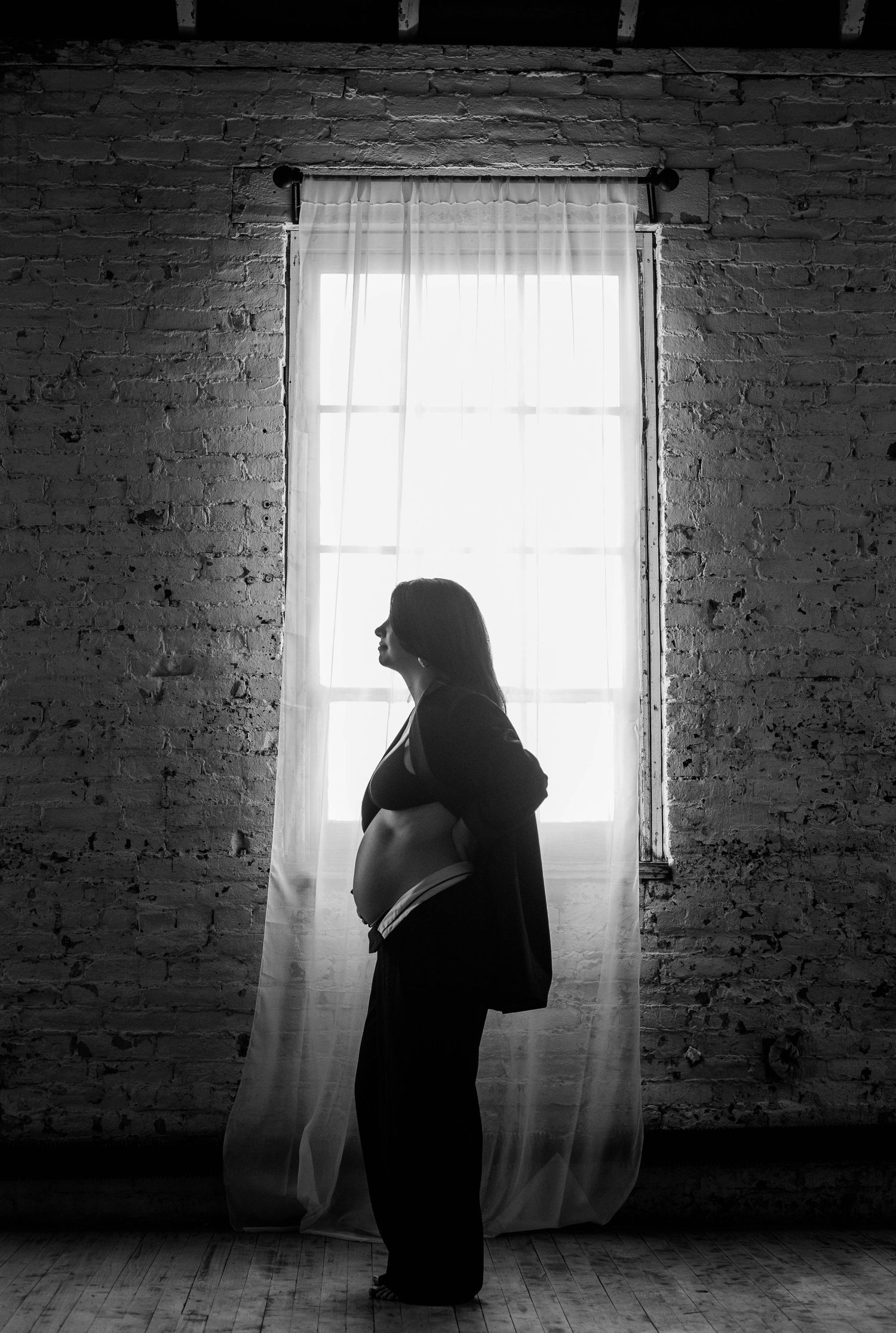 Black and white silhouette of a pregnant woman standing by a bright window in a brick-walled room.