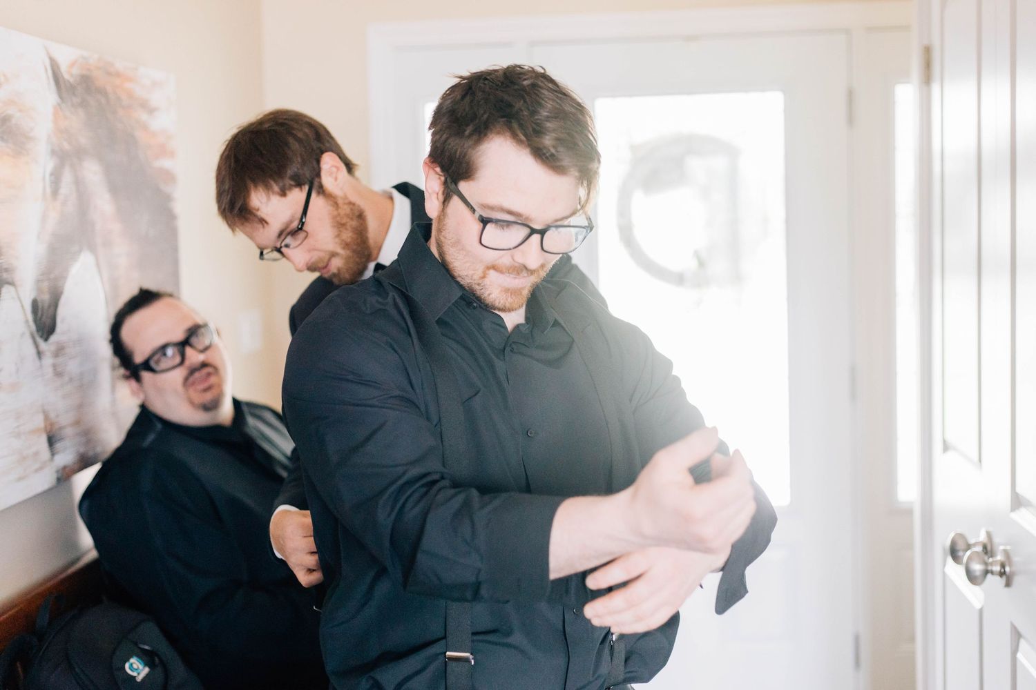 Three groomsmen in black suits laughing and helping each other get dressed before wedding.