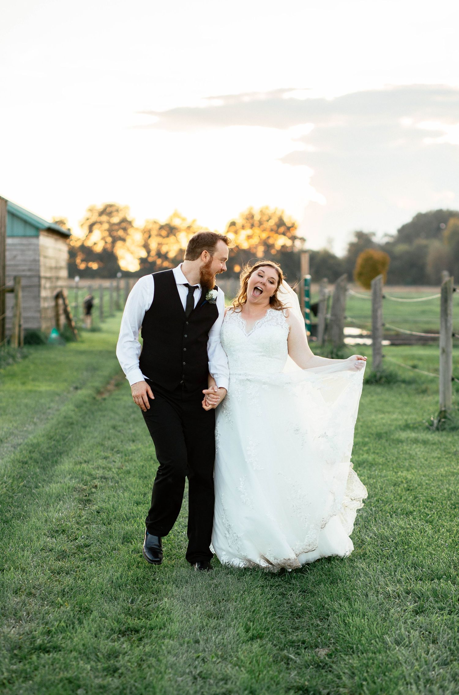 Laughing bride and groom strolling hand in hand across a sunlit farm at sunset.