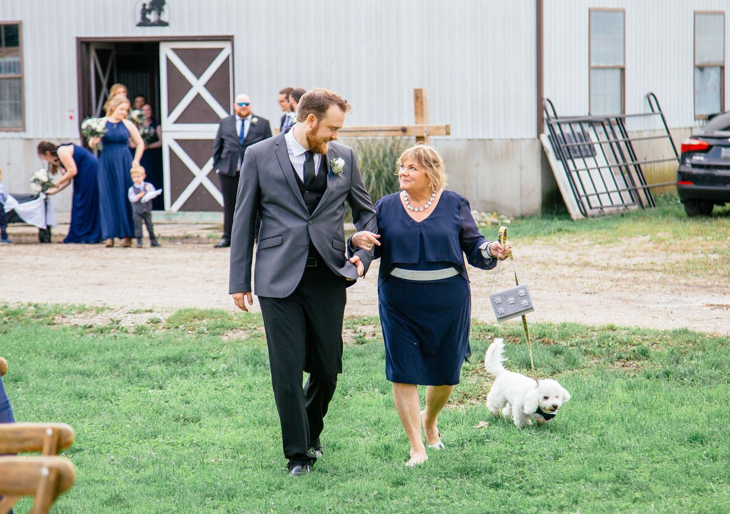 Groom in gray suit walks with a woman in navy dress and a small white dog carrying a sign at an outdoor wedding.