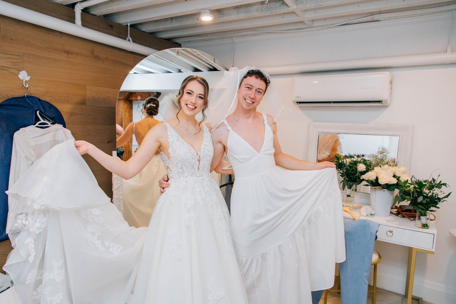 Two bridesmaids in white wedding dresses smile while holding skirts in bridal suite.