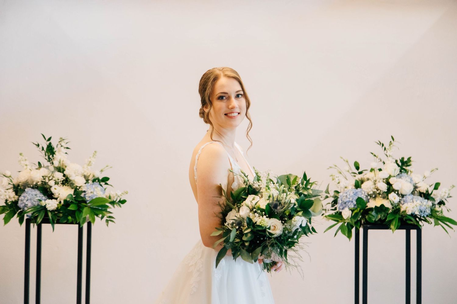 Elegant bride in sleeveless lace gown holding large white and green bouquet beside floral arrangements.