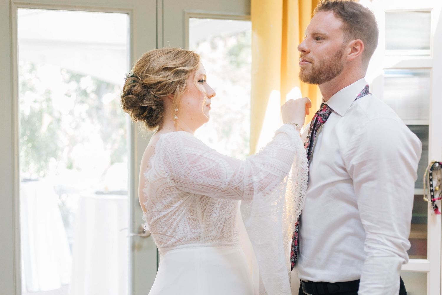 A bride in a lace dress adjusting the tie of a groom in a white shirt indoors.