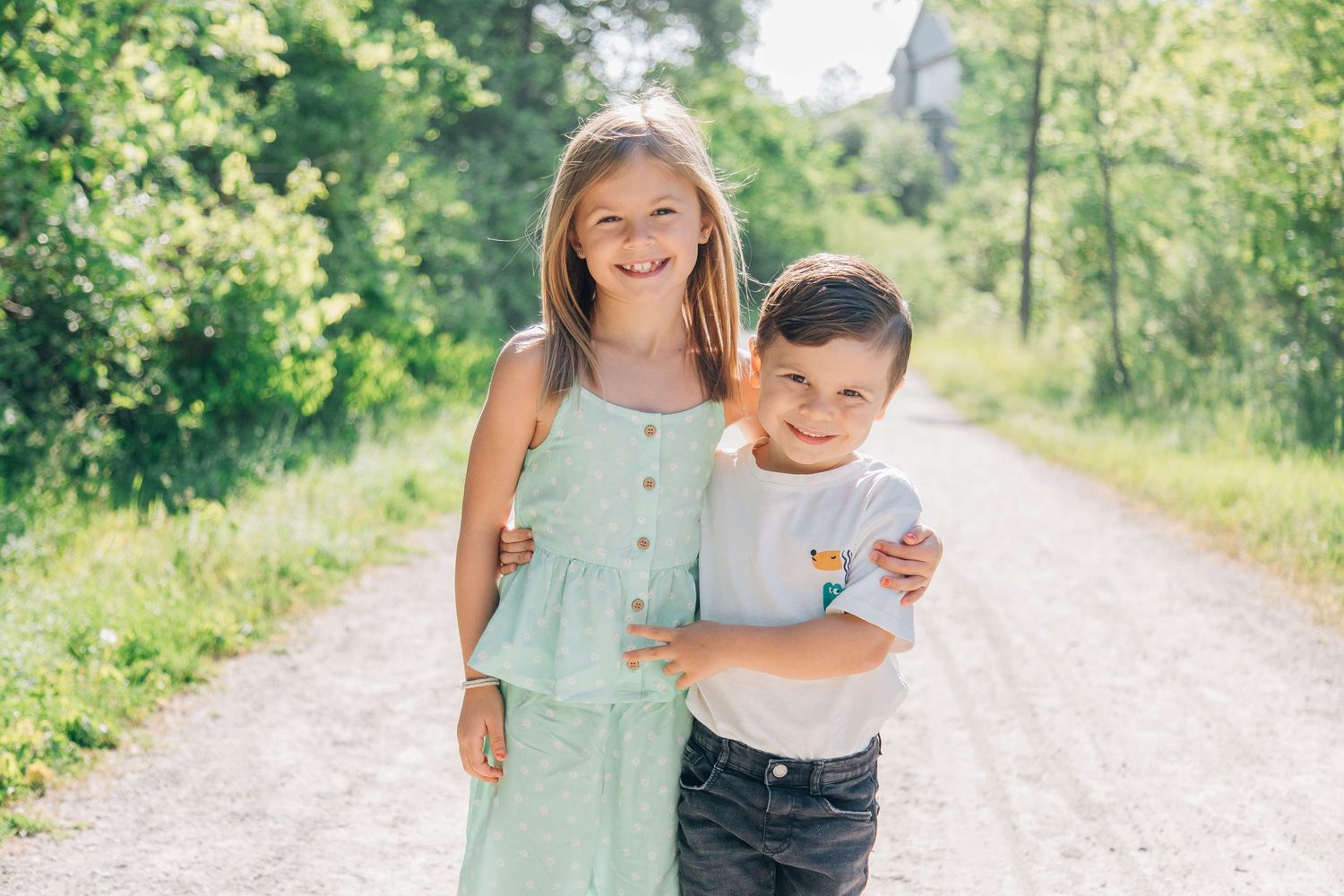 Smiling young girl in mint dress and little boy hugging on sunny gravel path.