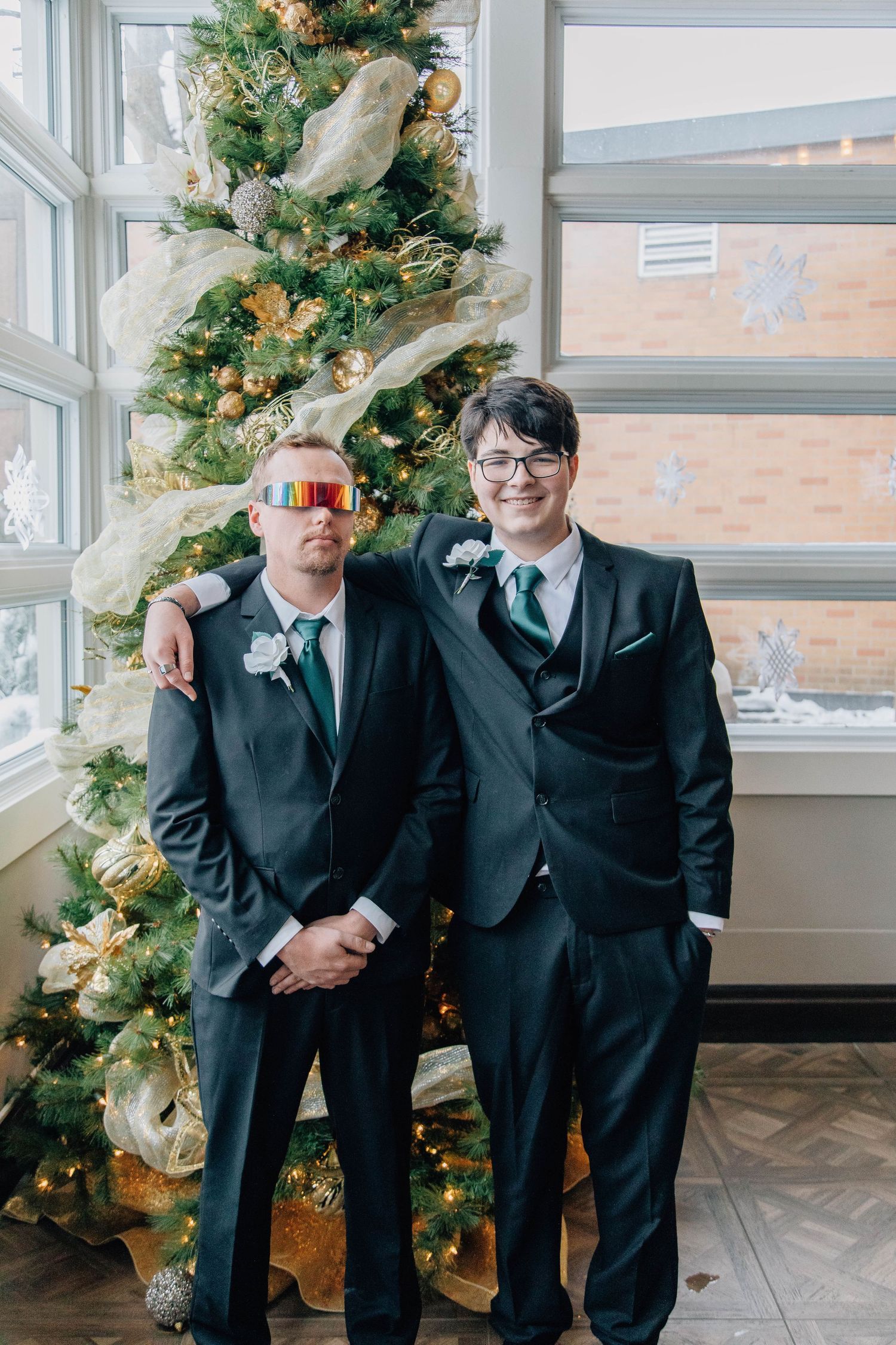 Two groomsmen in dark suits with green ties pose in front of decorated Christmas tree at winter wedding ceremony.