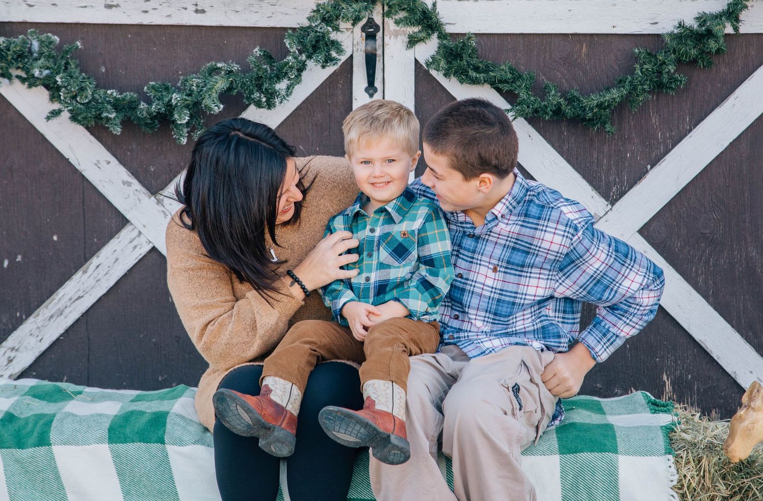 Parents kissing smiling toddler boy on cheeks during Christmas barn photo session.