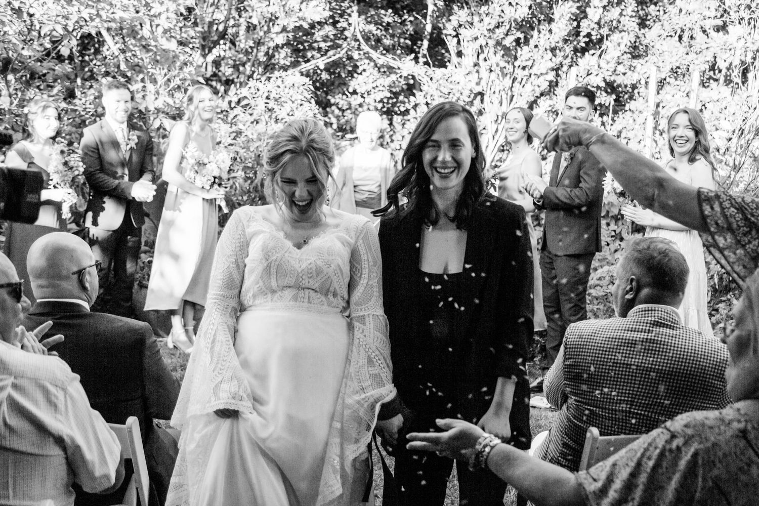 Two brides walk joyfully down aisle through confetti shower after outdoor garden wedding ceremony.