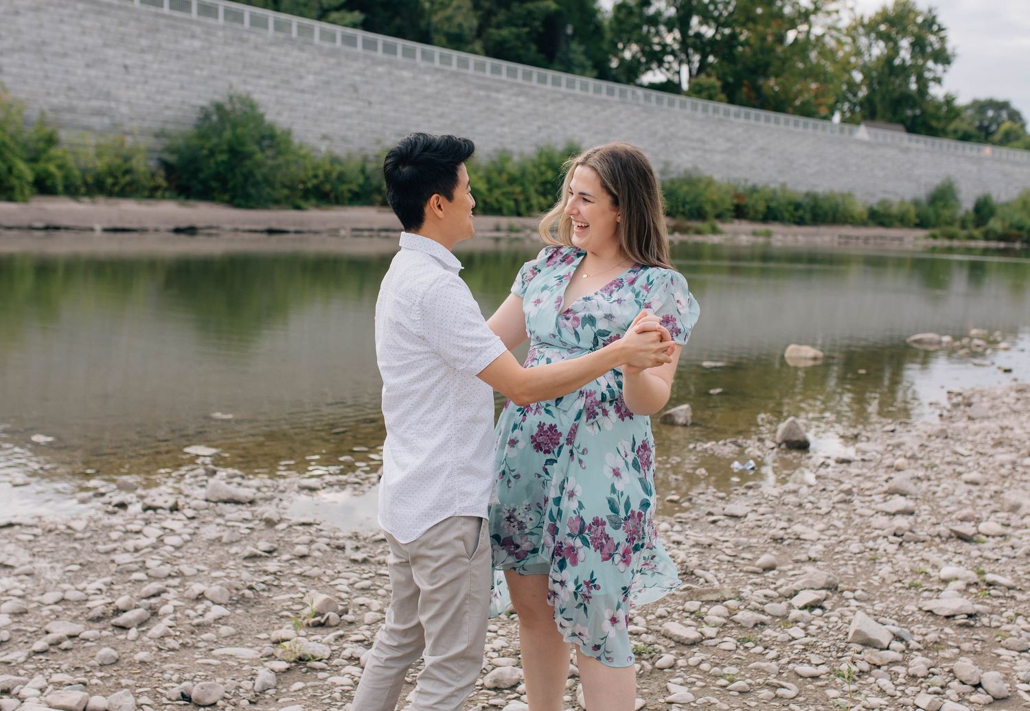 Couple laughing and embracing near a rocky riverbank with stone wall in background.
