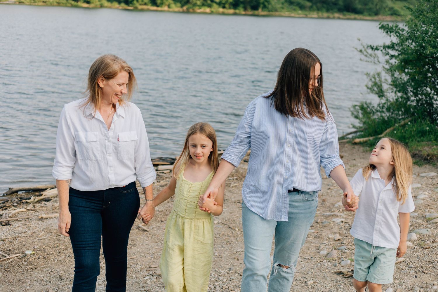 Two mothers holding hands with two young daughters walking along a rocky lakeside on a sunny day.