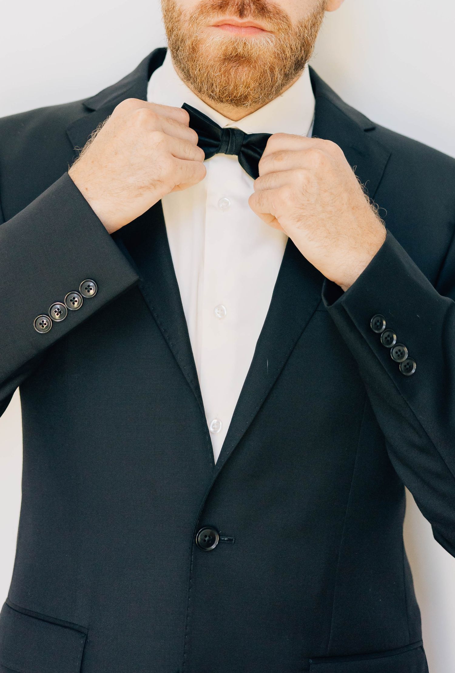 Groom in black tuxedo adjusting black bow tie against a white background.