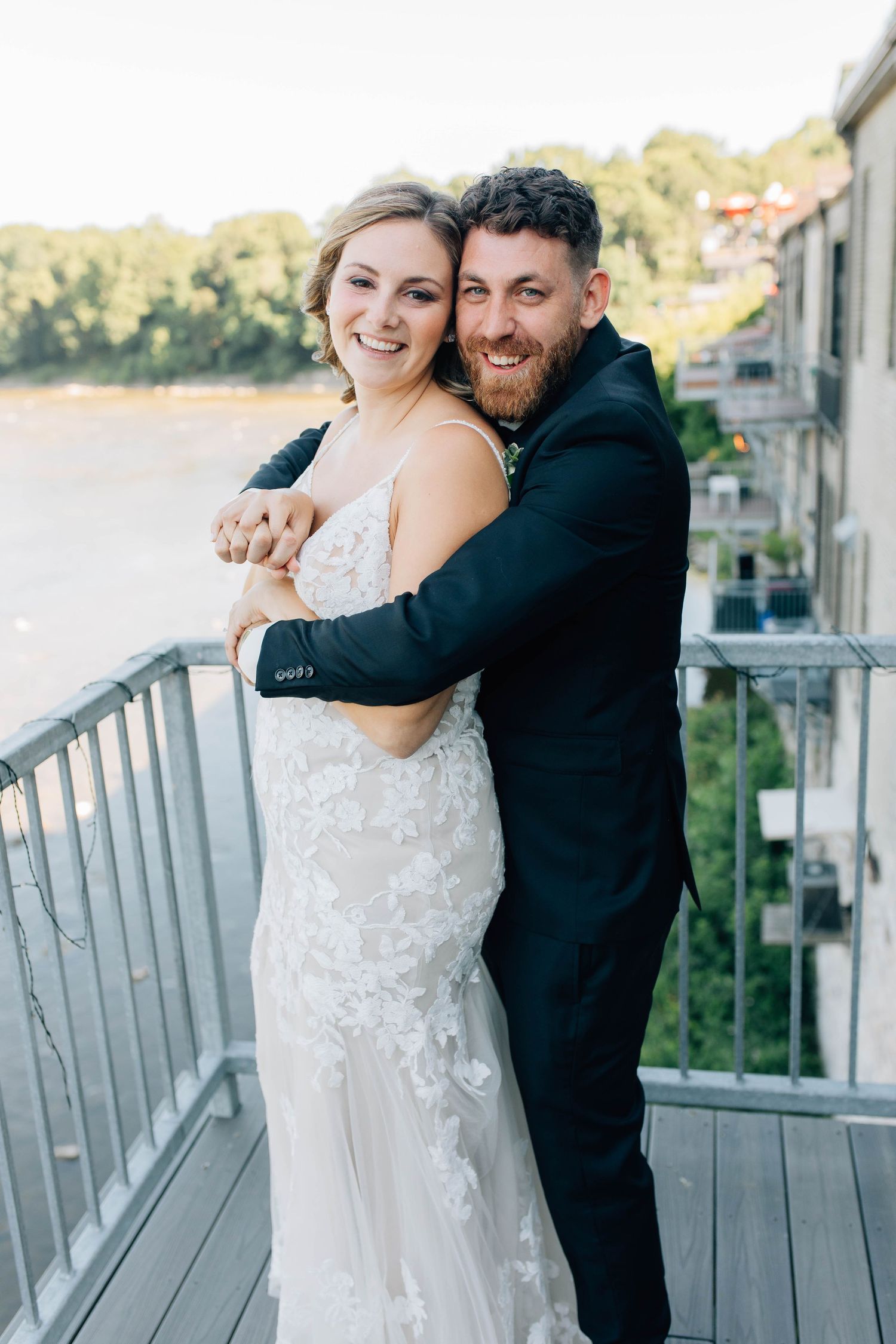 Smiling bride and bearded groom embrace on outdoor balcony with river and trees in background.