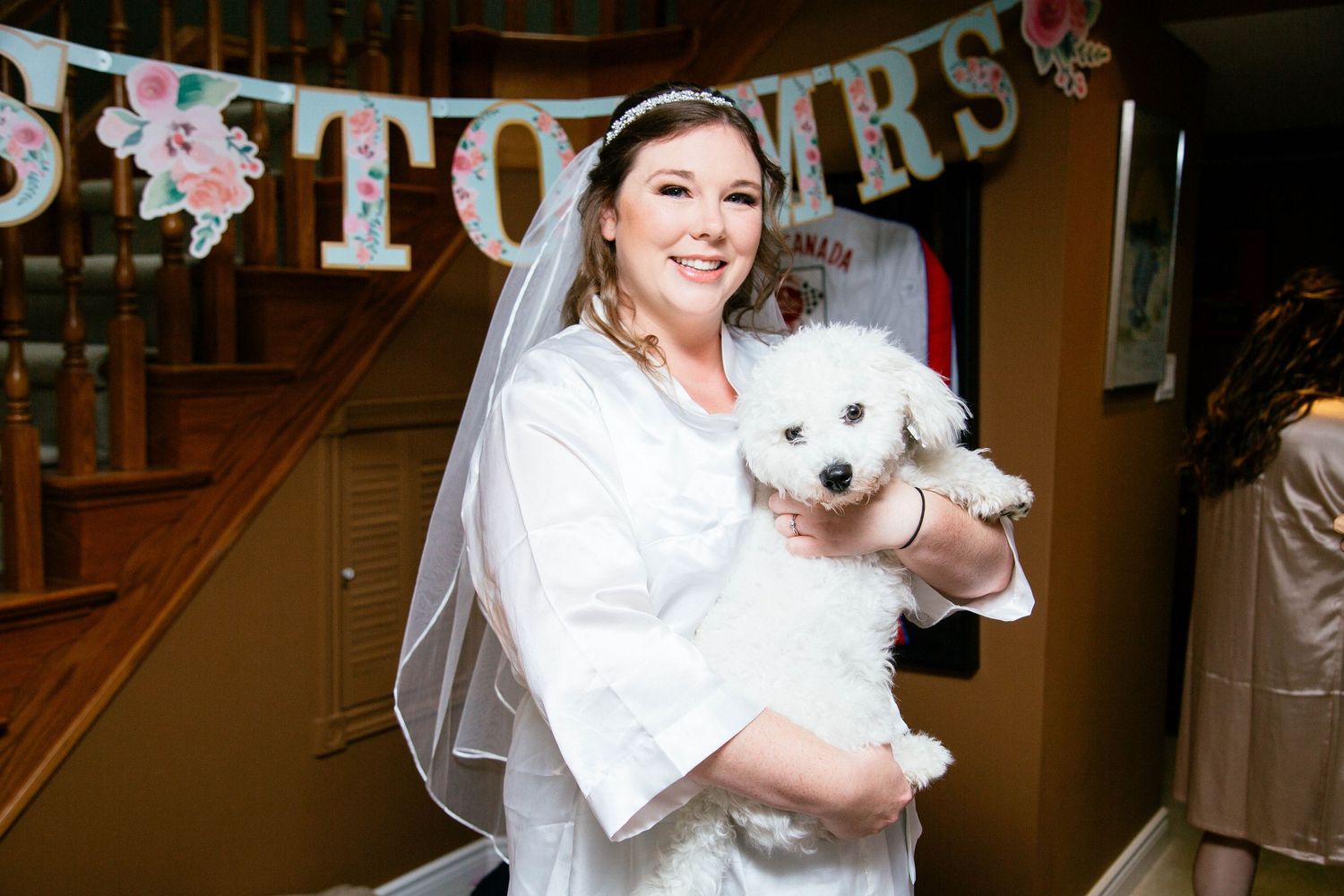Bride in white robe and veil holds a fluffy white dog near a MRS to MRS banner.