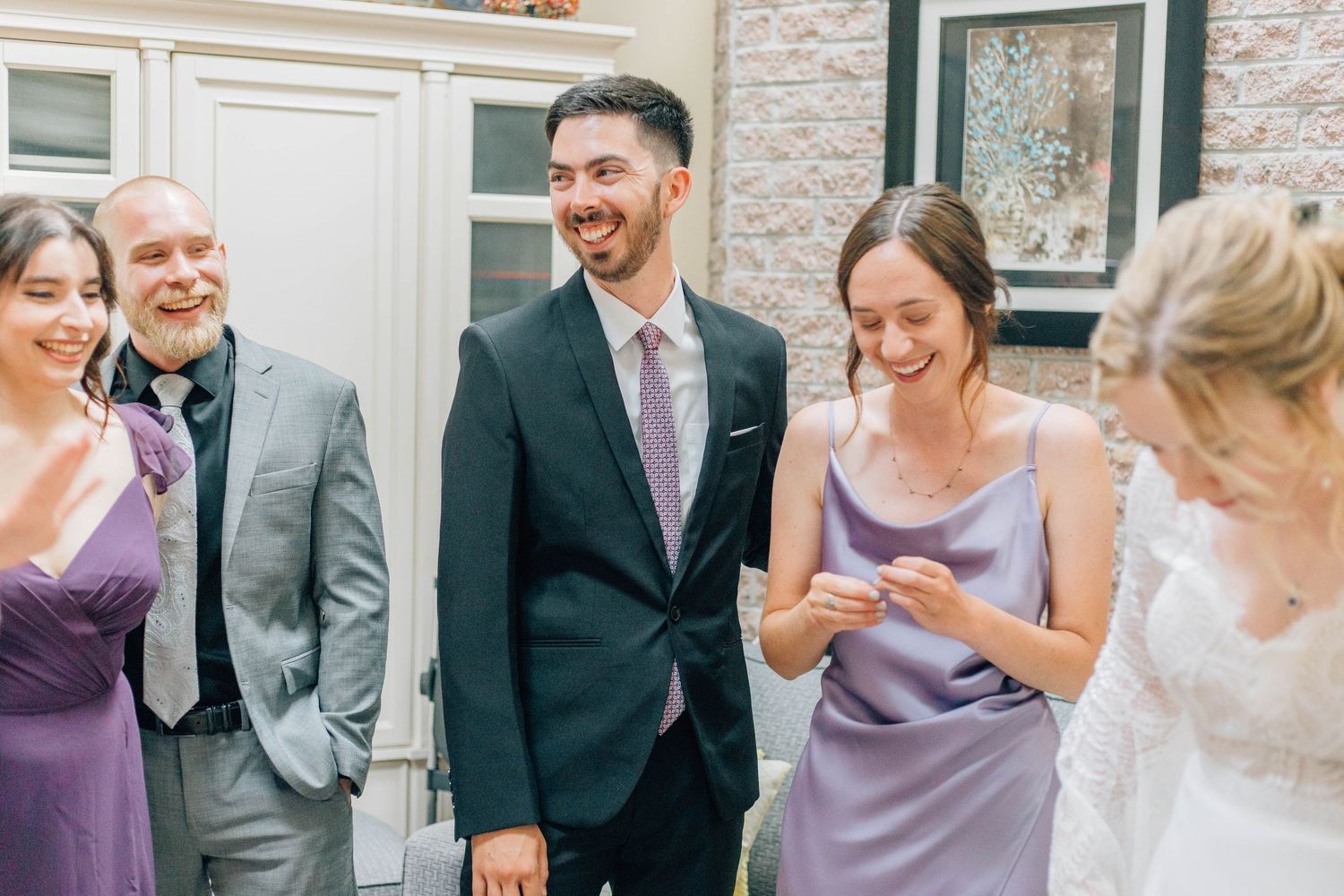 Smiling groom in dark suit stands with wedding guests in purple attire at reception.