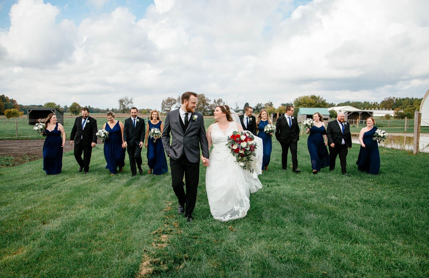 Bride and groom walk across green lawn with wedding party in navy blue dresses and gray suits on a cloudy day.