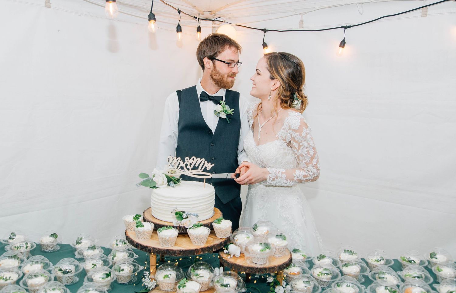 Newlyweds cutting their elegant white wedding cake surrounded by cupcakes at reception.