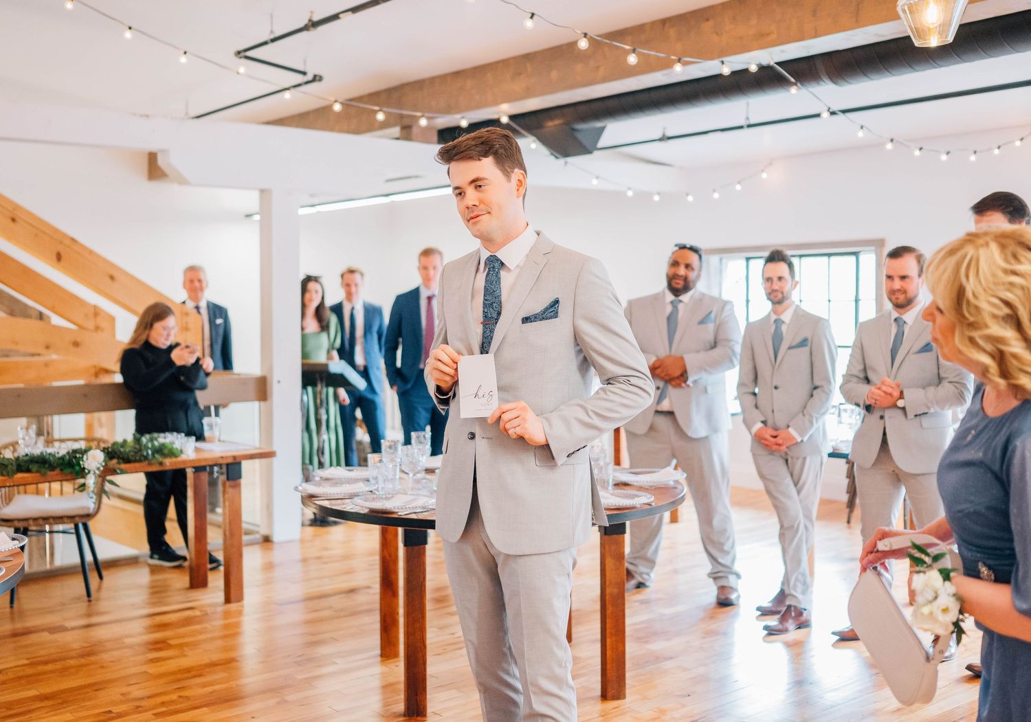 Groom in light gray suit speaks to wedding guests in elegant venue with string lights.