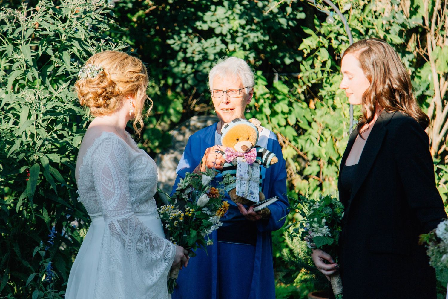 Wedding officiant holding teddy bear reads ceremony script between two brides in garden setting.