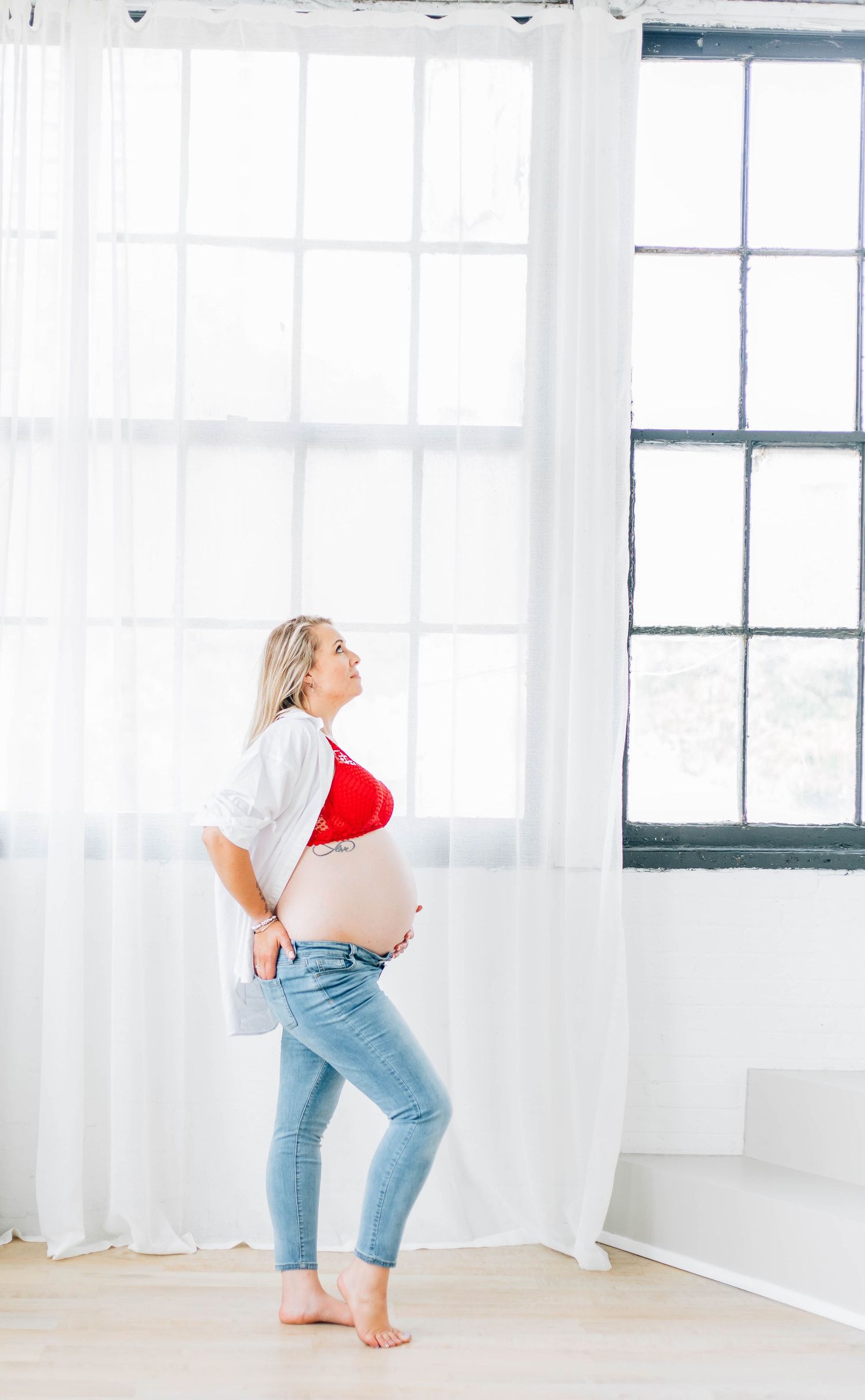 Pregnant woman in red bralette, white cardigan, and jeans standing by bright windows holding ultrasound photo.