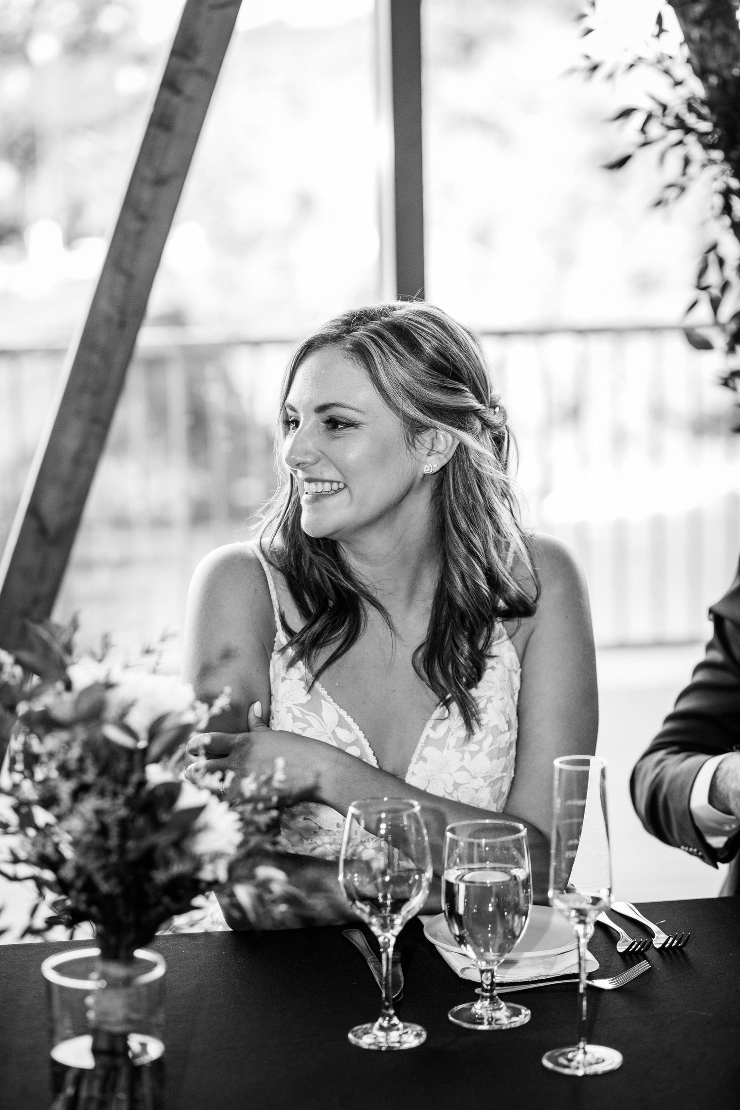 Black and white portrait of a smiling bride at the reception table with wine glasses and floral arrangements.