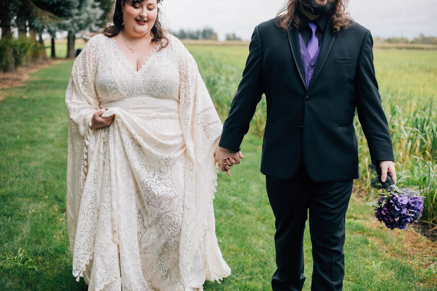 Bride in lace kimono gown and groom in black suit holding purple bouquet walking through vineyard rows.