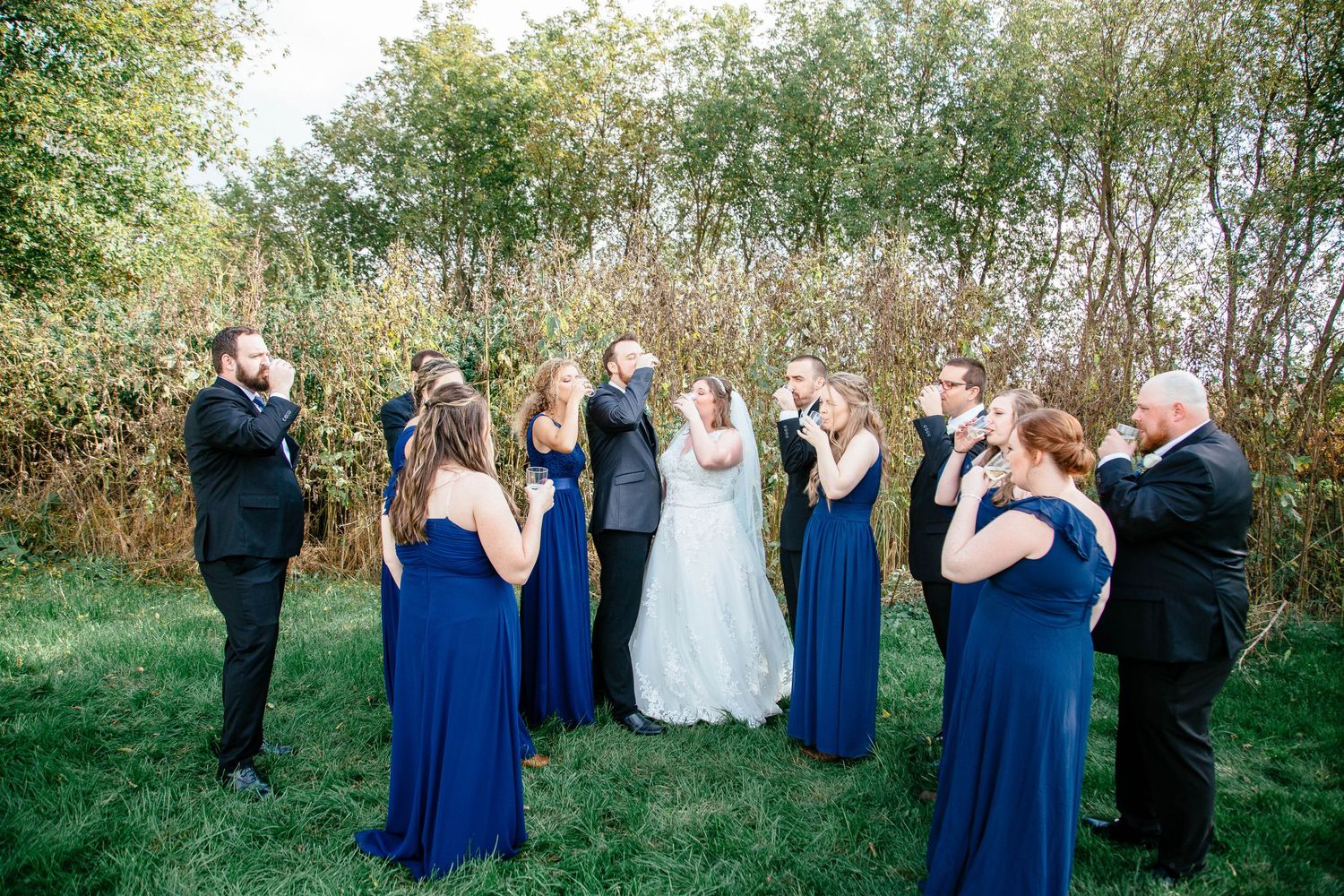 Wedding party celebrates outdoors, bridesmaids in navy blue gowns and groomsmen in black suits cheering for bride and groom.
