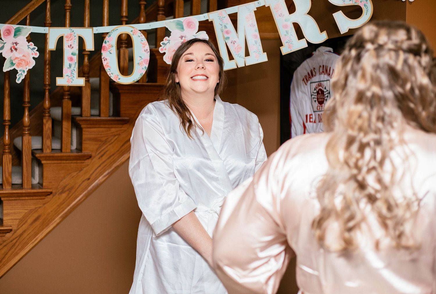 Bride in white robe smiles at bridesmaid near staircase decorated with MRS to MRS banner.