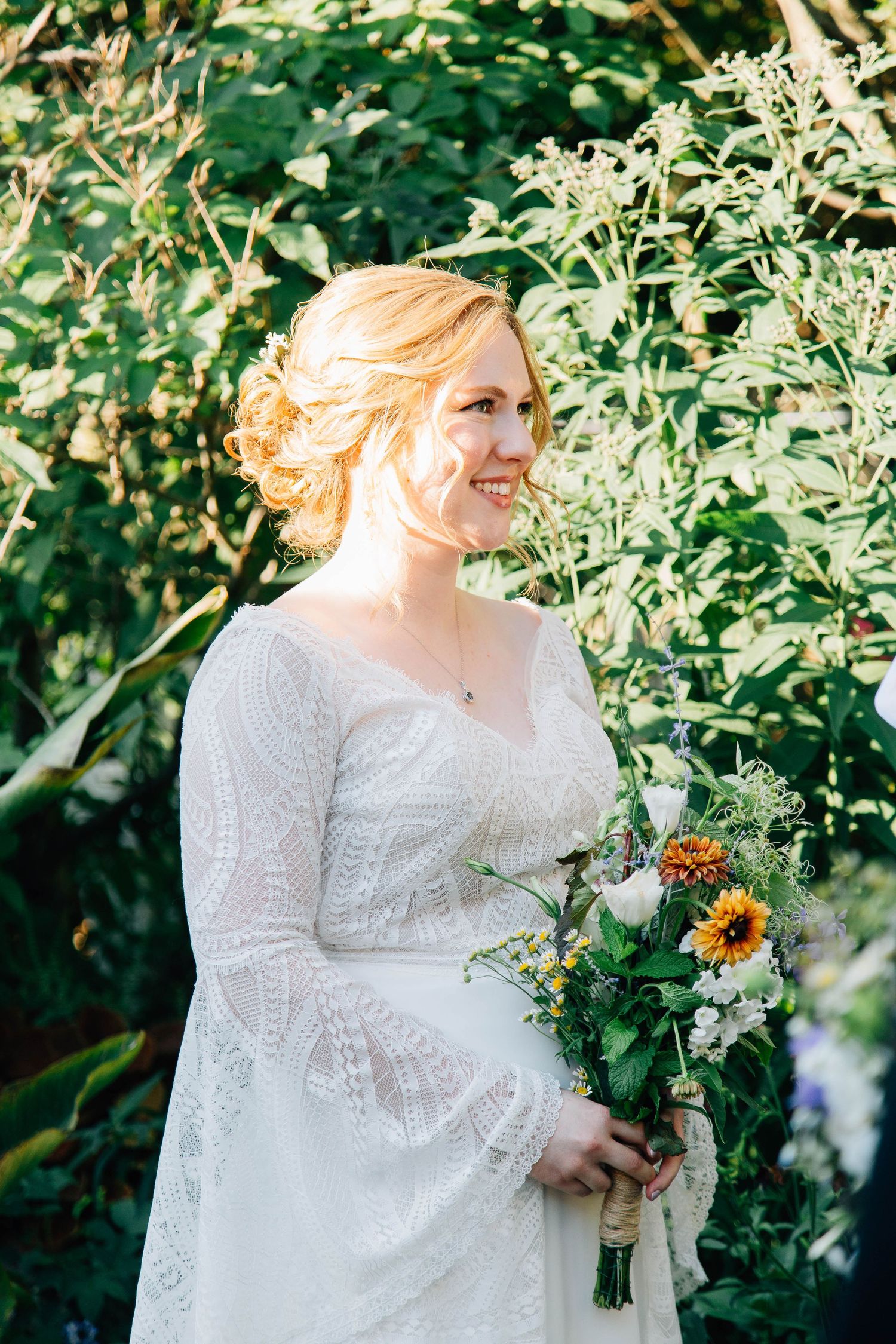 Smiling bride in lace long-sleeve wedding dress holding sunflower bouquet outdoors in lush garden.