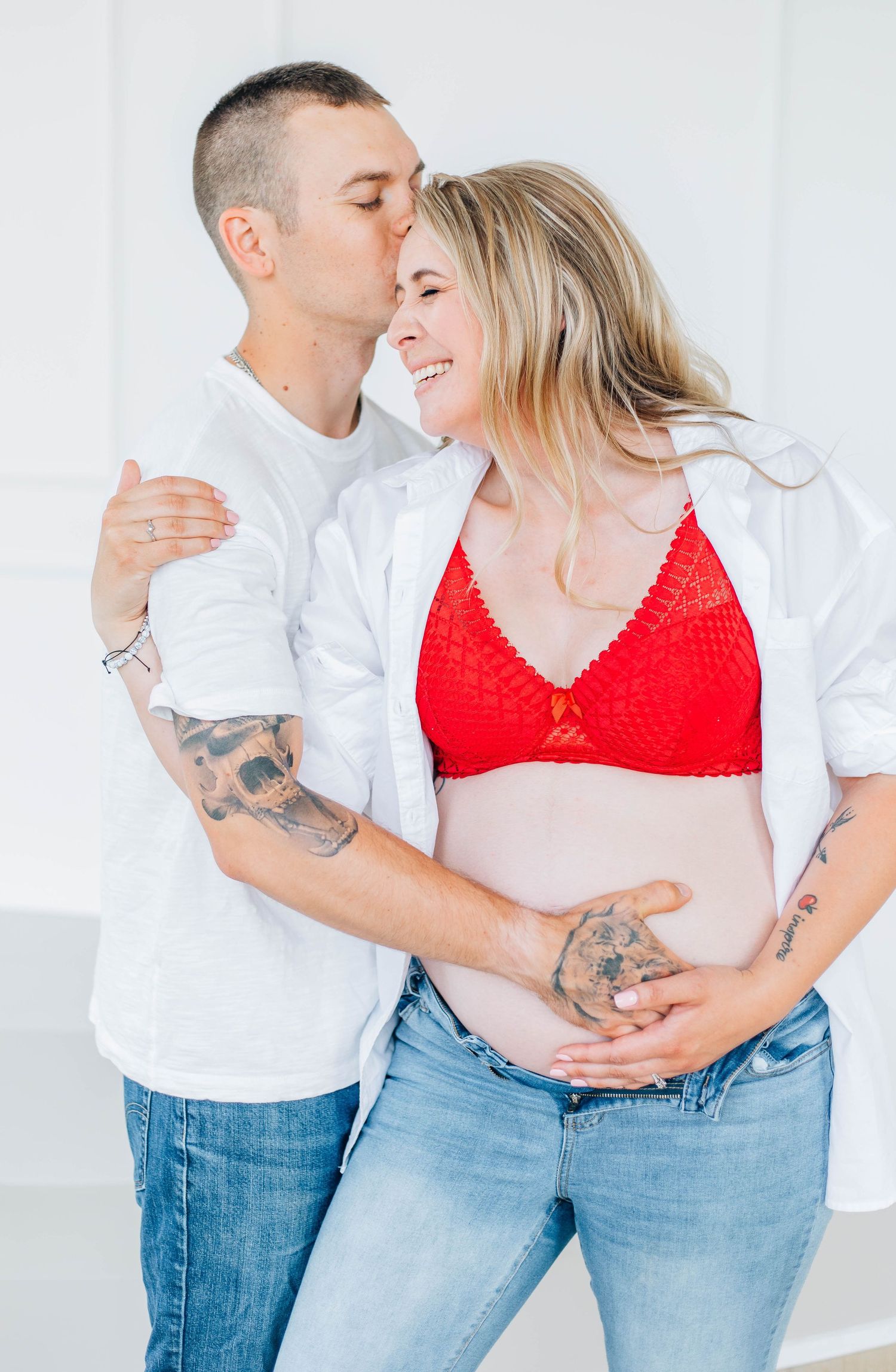 Man kissing pregnant woman's cheek as she smiles in red bralette and jeans.