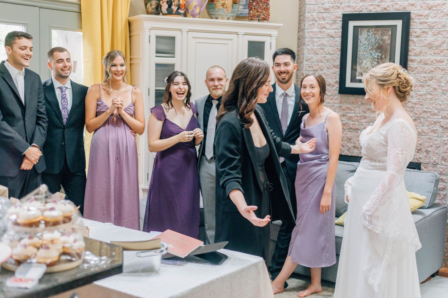 Bride in white lace gown meets cheerful wedding guests in purple dresses at reception.